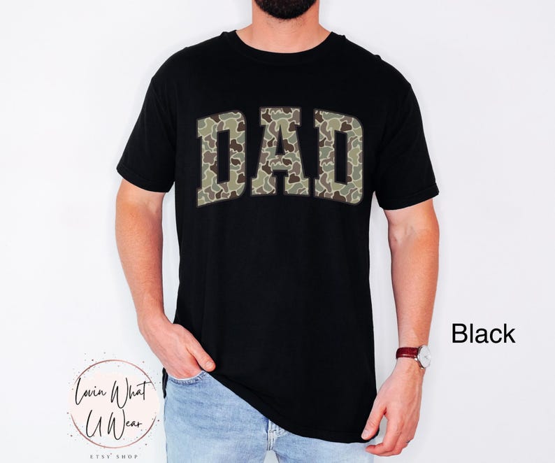 May include: Black crew neck t-shirt with the word "DAD" in a camouflage pattern across the chest. The shirt is worn by a person. The text "Black" is visible in the image.