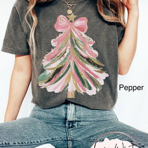 Op de afbeelding: Een donkergrijs t-shirt met een kerstboomontwerp. De boom is gemaakt van roze, groene en gouden penseelstreken, bekroond met een roze strik en een ster. Het woord "Pepper" staat op het shirt.