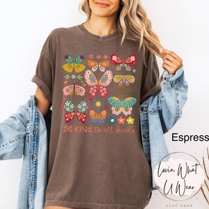 May include: A brown t-shirt with a graphic of colorful butterflies and flowers. The text "Be Kind to all kinds" is printed below the graphic.