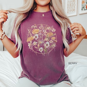 May include: A purple t-shirt featuring a circular floral design. The flowers are yellow, white, and brown. The text "BERRY" is in the bottom right corner of the image.