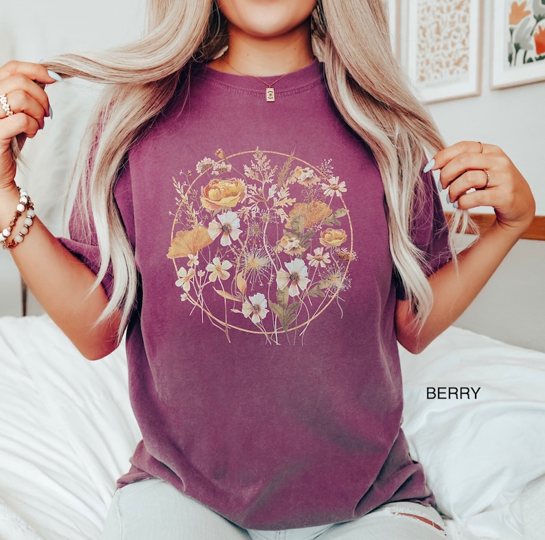 May include: A purple t-shirt featuring a circular floral design. The flowers are yellow, white, and brown. The text "BERRY" is in the bottom right corner of the image.