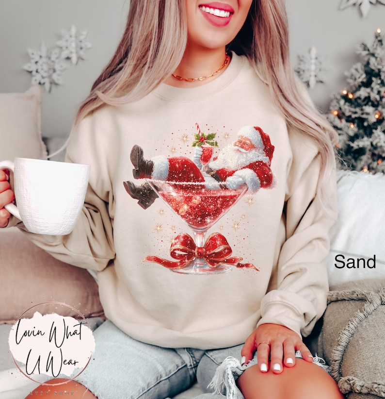 May include: Sand-colored sweatshirt featuring a graphic of Santa Claus relaxing in a cocktail glass. The design includes a red bow, glitter, and the text "Lovin What U Wear". A festive and fun holiday-themed apparel item.