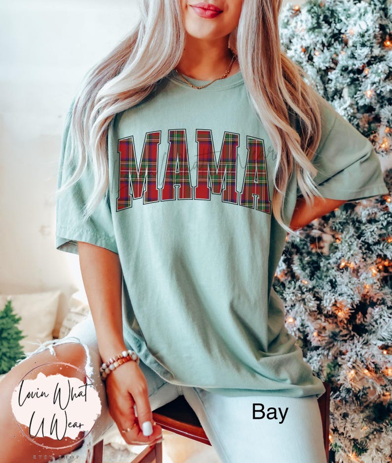 May include: A light green t-shirt with the word "MAMA" in large, red and green plaid letters across the front. The shirt is a relaxed fit and has short sleeves. The text "Bay" is at the bottom right of the image.
