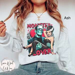 Peut inclure: Un sweat-shirt gris avec un imprim&eacute; graphique de Frankenstein et une femme en robe noire. Le graphique dit "Monster Lover" et pr&eacute;sente un c&oelig;ur rouge.
