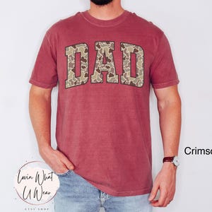 May include: Crimson-colored t-shirt with the word "DAD" in a camouflage pattern across the chest. The shirt has short sleeves and a crew neck. The text "Crimson" is visible in the lower right corner.