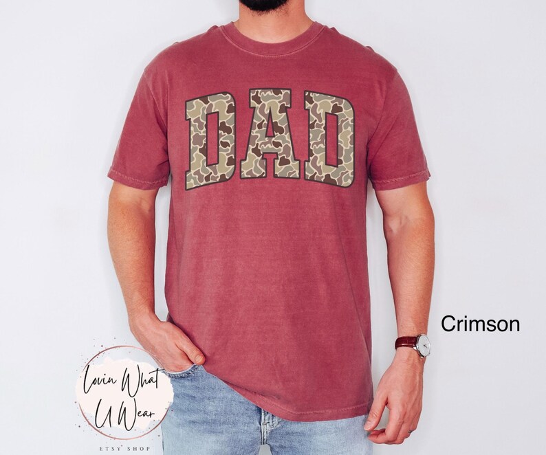 May include: Crimson-colored t-shirt with the word "DAD" in a camouflage pattern across the chest. The shirt has short sleeves and a crew neck. The text "Crimson" is visible in the lower right corner.