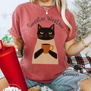 May include: Crimson-colored t-shirt with the text "Sweater Weather" above a black cat graphic holding a mug. The cat is wearing a striped sweater. The shirt is a comfortable, casual style.