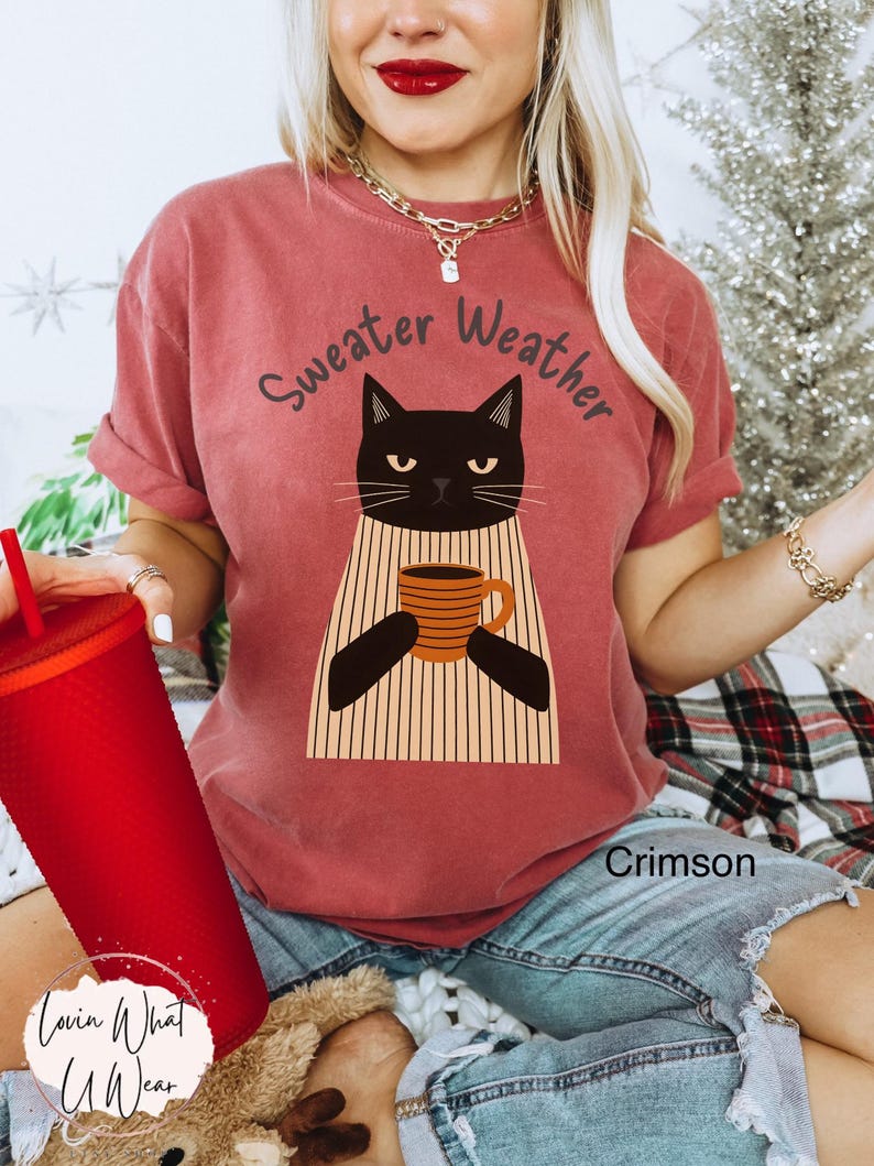 May include: Crimson-colored t-shirt with the text "Sweater Weather" above a black cat graphic holding a mug. The cat is wearing a striped sweater. The shirt is a comfortable, casual style.