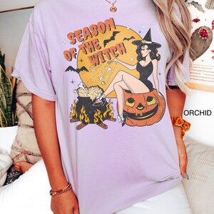 Puede incluir: Una camiseta lavanda con un gr&aacute;fico vintage de una bruja sentada en una calabaza. El gr&aacute;fico dice "Season of the Witch" y presenta murci&eacute;lagos, un caldero y huesos.