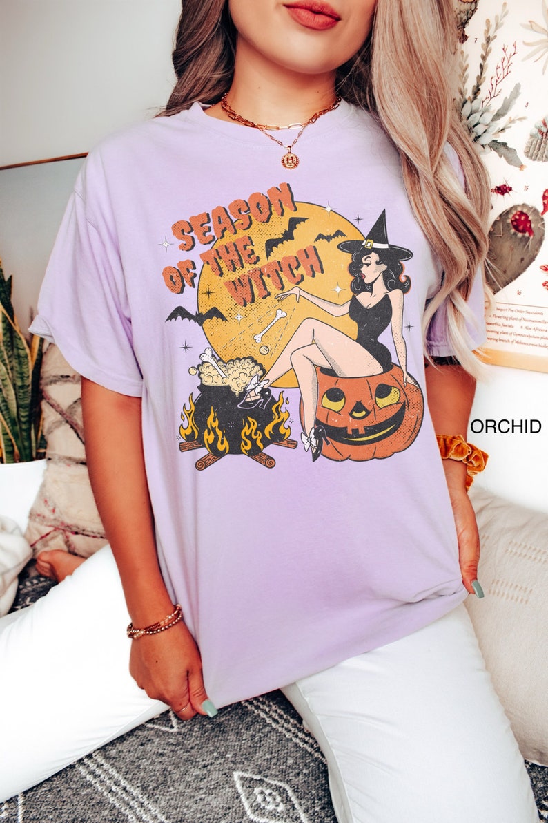 Puede incluir: Una camiseta lavanda con un gr&aacute;fico vintage de una bruja sentada en una calabaza. El gr&aacute;fico dice "Season of the Witch" y presenta murci&eacute;lagos, un caldero y huesos.