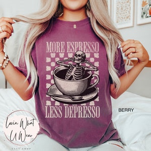 May include: A purple t-shirt with a graphic of a skeleton relaxing in a cup of coffee. The text "More Espresso Less Depresso" is printed on the shirt.