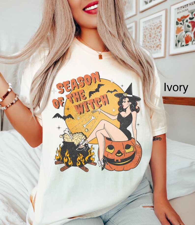 Puede incluir: Una camiseta blanca con una ilustraci&oacute;n de estilo vintage de una bruja sentada sobre una calabaza. El texto "Season of the Witch" est&aacute; impreso en la camiseta. La camiseta est&aacute; disponible en marfil.