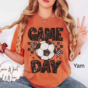 May include: Orange t-shirt with the words "GAME DAY" in a distressed black font, a soccer ball graphic, and lightning bolt and checkered flag accents. The shirt is a comfortable fit.