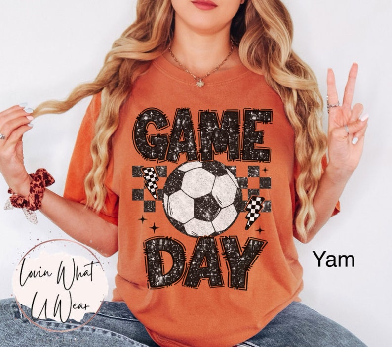 May include: Orange t-shirt with the words "GAME DAY" in a distressed black font, a soccer ball graphic, and lightning bolt and checkered flag accents. The shirt is a comfortable fit.
