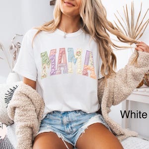 Puede incluir: Camiseta blanca con la palabra "MAMA" en letras coloridas de estilo patchwork. Las letras tienen dise&ntilde;os florales y estampados. La modelo lleva pantalones cortos de mezclilla y una chaqueta beige. El texto "White" es visible en la parte inferior derecha.