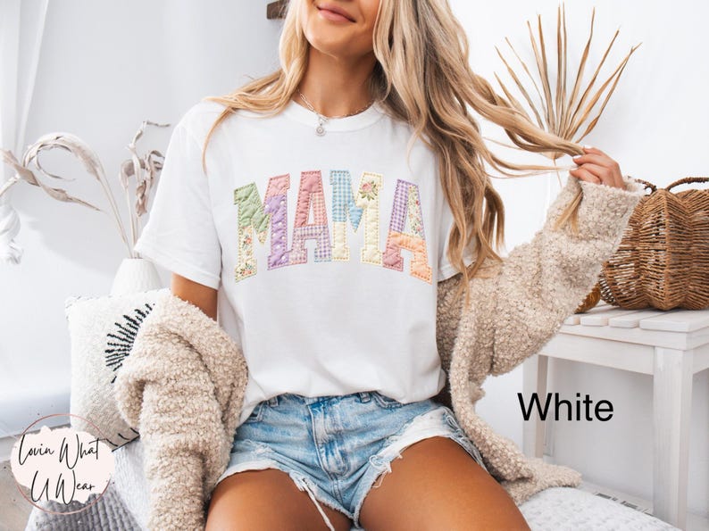 Puede incluir: Camiseta blanca con la palabra "MAMA" en letras coloridas de estilo patchwork. Las letras tienen dise&ntilde;os florales y estampados. La modelo lleva pantalones cortos de mezclilla y una chaqueta beige. El texto "White" es visible en la parte inferior derecha.