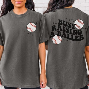 May include: A grey t-shirt with a baseball graphic on the front and the text "Busy Raising A Baller" with a baseball graphic on the back.