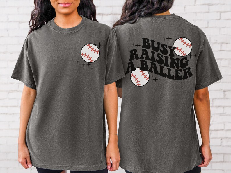 May include: A grey t-shirt with a baseball graphic on the front and the text "Busy Raising A Baller" with a baseball graphic on the back.