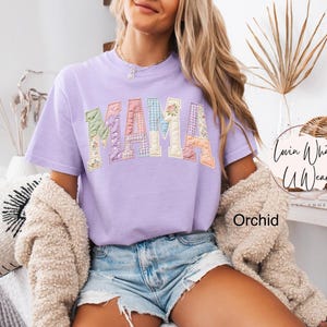 Puede incluir: Una camiseta lila con la palabra "MAMA" en letras de patchwork. Las letras est&aacute;n hechas de varias telas pastel, incluyendo estampados florales y de cuadros. Es una prenda de vestir.
