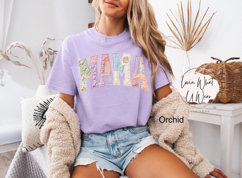Puede incluir: Una camiseta lila con la palabra "MAMA" en letras de patchwork. Las letras est&aacute;n hechas de varias telas pastel, incluyendo estampados florales y de cuadros. Es una prenda de vestir.
