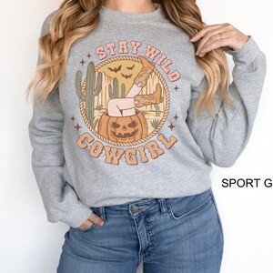 May include: A grey sweatshirt with a graphic print of a woman in cowboy boots sitting in a jack-o-lantern. The text "Stay Wild Cowgirl" is printed around the graphic. The sweatshirt is labelled "Sport Grey".
