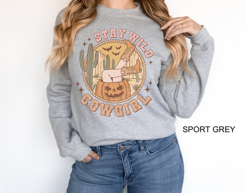 May include: A grey sweatshirt with a graphic print of a woman in cowboy boots sitting in a jack-o-lantern. The text "Stay Wild Cowgirl" is printed around the graphic. The sweatshirt is labelled "Sport Grey".