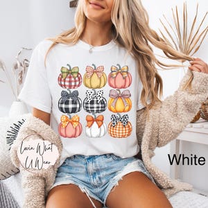 Peut inclure: T-shirt blanc avec un motif de citrouilles color&eacute;es. Les citrouilles pr&eacute;sentent diff&eacute;rents motifs et n&oelig;uds, notamment des carreaux et des pois. Le texte "Lovin What U Wear" est visible sur le t-shirt.