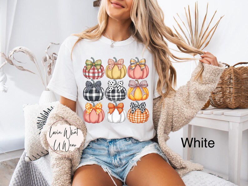 Peut inclure: T-shirt blanc avec un motif de citrouilles color&eacute;es. Les citrouilles pr&eacute;sentent diff&eacute;rents motifs et n&oelig;uds, notamment des carreaux et des pois. Le texte "Lovin What U Wear" est visible sur le t-shirt.