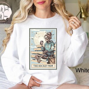 May include: White sweatshirt featuring a graphic of a skeleton holding a coffee cup and hockey stick. The graphic is in a vintage style with a blue and brown colour scheme. The text "The Hockey Mom" is below the graphic.