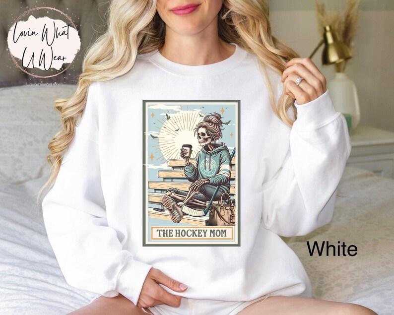 May include: White sweatshirt featuring a graphic of a skeleton holding a coffee cup and hockey stick. The graphic is in a vintage style with a blue and brown colour scheme. The text "The Hockey Mom" is below the graphic.
