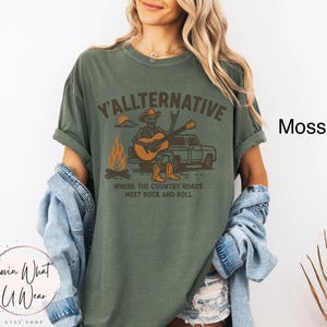 May include: Moss green t-shirt with the text "Y'ALLTERNATIVE" and a graphic of a skeleton playing a guitar next to a campfire and a truck. The text "WHERE THE COUNTRY ROADS MEET ROCK AND ROLL" is below the graphic. The shirt is from the Lovin What U Wear Etsy shop.