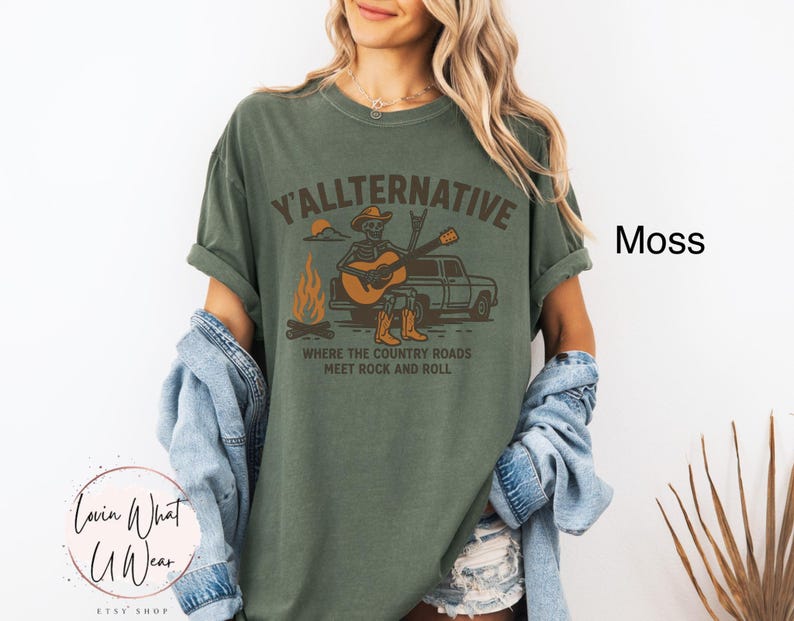 May include: Moss green t-shirt with the text "Y'ALLTERNATIVE" and a graphic of a skeleton playing a guitar next to a campfire and a truck. The text "WHERE THE COUNTRY ROADS MEET ROCK AND ROLL" is below the graphic. The shirt is from the Lovin What U Wear Etsy shop.