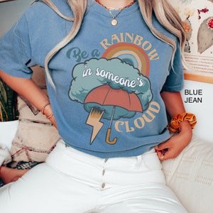 May include: A blue t-shirt with a graphic of a rainbow, a cloud, an umbrella, and lightning bolt. The text on the shirt reads "Be a rainbow in someone's cloud".