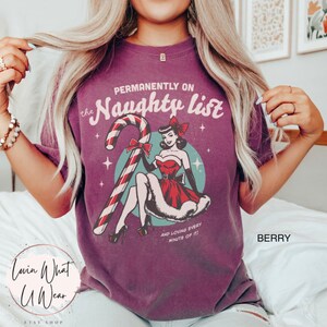 May include: Purple t-shirt with a vintage-style illustration of a woman in a red dress holding a candy cane. The text "Permanently on the Naughty List" is printed above the illustration, and the text "And loving every minute of it!" is printed below the illustration.