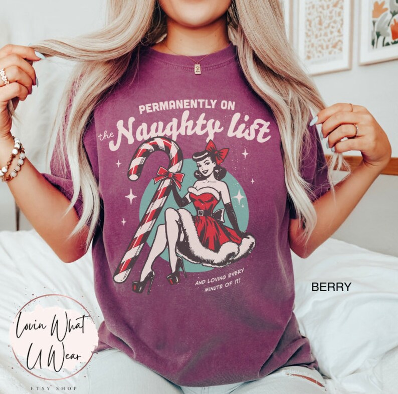 May include: Purple t-shirt with a vintage-style illustration of a woman in a red dress holding a candy cane. The text "Permanently on the Naughty List" is printed above the illustration, and the text "And loving every minute of it!" is printed below the illustration.