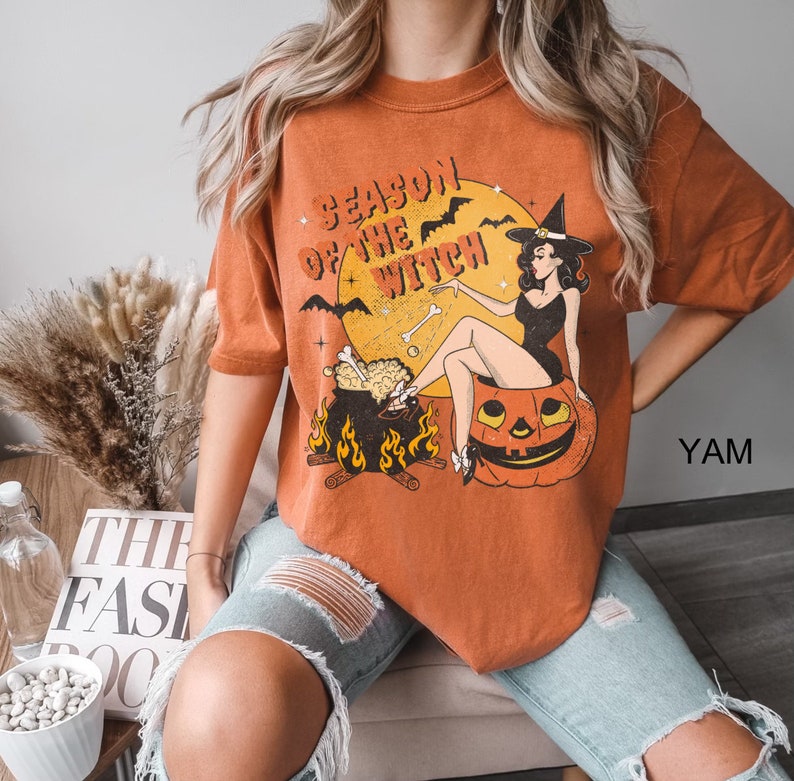 Puede incluir: Camiseta naranja con una ilustraci&oacute;n vintage de una bruja sentada en una calabaza. La bruja lleva un vestido negro y un sombrero puntiagudo. El texto de la camiseta dice "Season of the Witch".