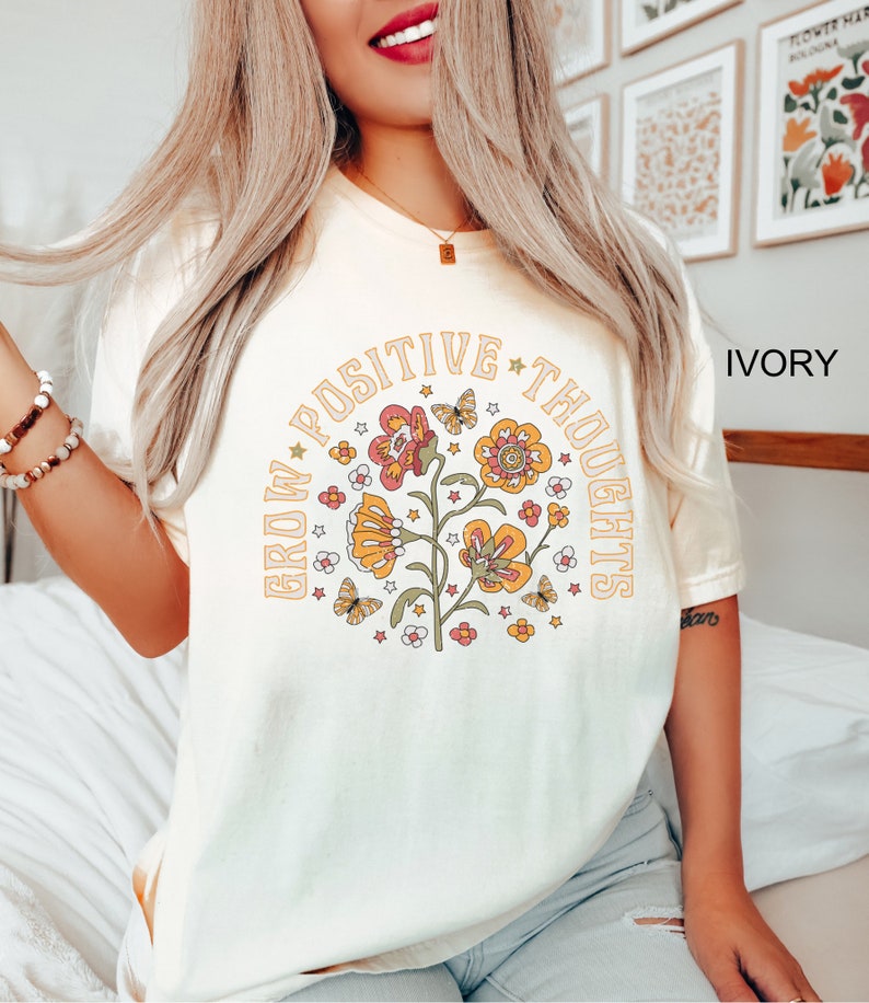 Op de afbeelding: Ivoorwit T-shirt met een afbeelding van bloemen en vlinders in een cirkel met de tekst "Grow Positive Thoughts".