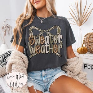 May include: A dark gray t-shirt with the words "Sweater Weather" in a decorative font, accented by a camouflage bow. The shirt is part of a fall-themed collection, perfect for casual wear.