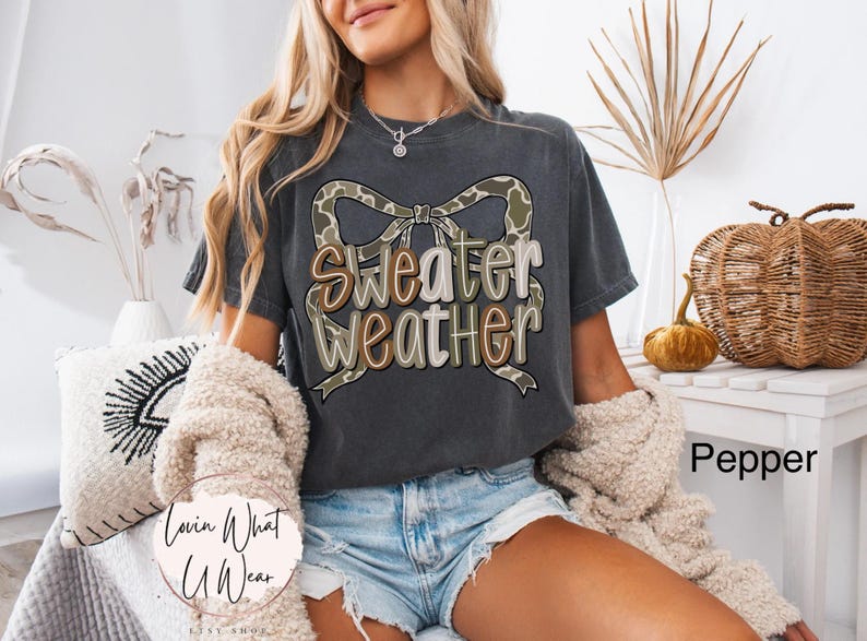 May include: A dark gray t-shirt with the words "Sweater Weather" in a decorative font, accented by a camouflage bow. The shirt is part of a fall-themed collection, perfect for casual wear.