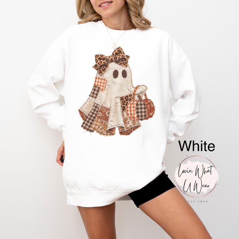Op de afbeelding: Een wit sweatshirt met lange mouwen en een centrale afbeelding van een geest versierd met een luipaardprint strik en patchworkpatronen in bruine en oranje tinten. De geest houdt een kleine pompoen vast. De tekst White en Lovin What U Wear is zichtbaar.