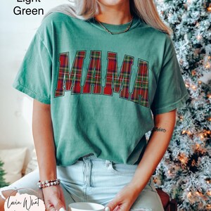 May include: Light green t-shirt with the word "MAMA" in red and green plaid letters. The shirt is a relaxed fit and has short sleeves. The image also shows a person wearing the shirt.