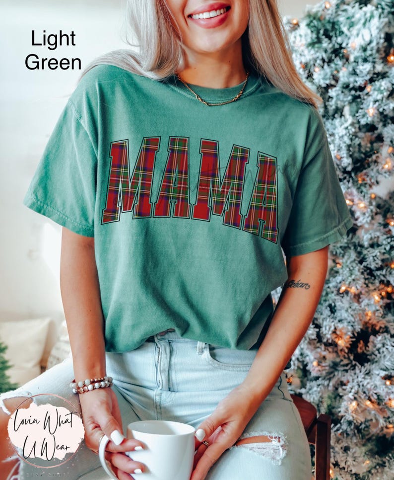 May include: Light green t-shirt with the word "MAMA" in red and green plaid letters. The shirt is a relaxed fit and has short sleeves. The image also shows a person wearing the shirt.