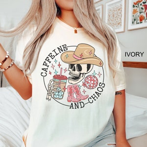 Pu&ograve; includere: Una maglietta bianca con una grafica di uno scheletro che indossa un cappello da cowboy, tiene una tazza di caff&egrave; e una palla da discoteca. Il testo "Caffeine and Chaos" &egrave; scritto intorno alla grafica.