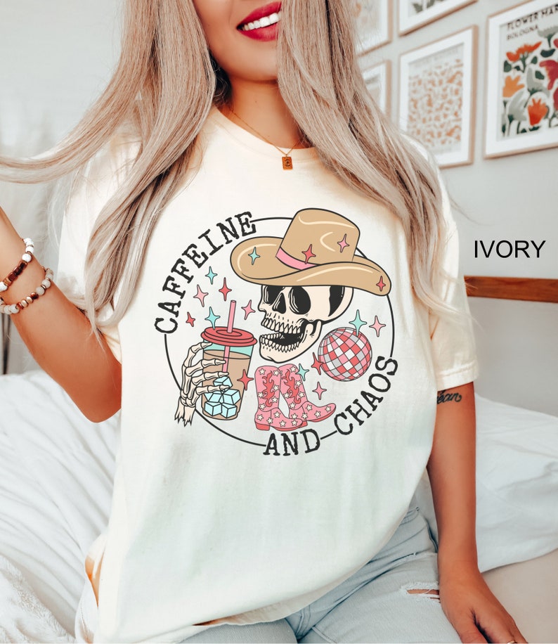 Pu&ograve; includere: Una maglietta bianca con una grafica di uno scheletro che indossa un cappello da cowboy, tiene una tazza di caff&egrave; e una palla da discoteca. Il testo "Caffeine and Chaos" &egrave; scritto intorno alla grafica.