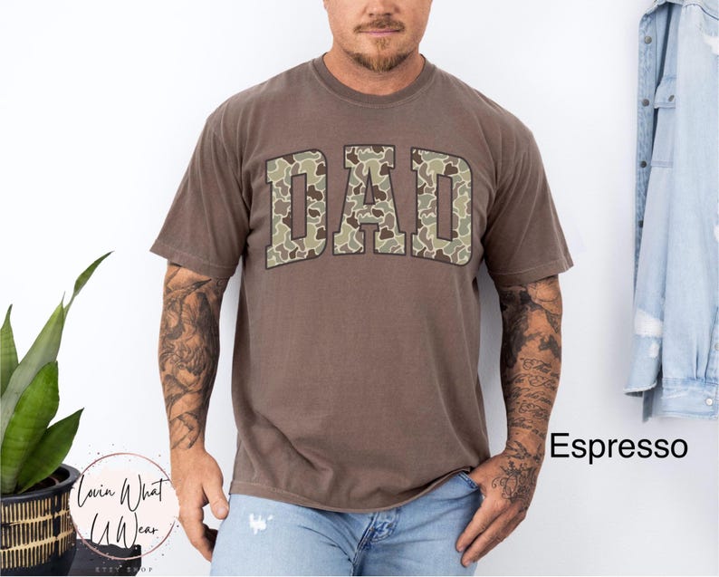 May include: Espresso-colored t-shirt with the word "DAD" in a camouflage pattern across the chest. The shirt has short sleeves and a crew neck. The model is wearing blue jeans. The shirt is part of the Lovin What U Wear Etsy shop.