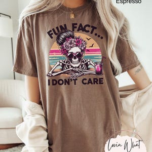May include: Brown t-shirt with a graphic of a skeleton with a flower crown and sunglasses holding a glass of pink liquid. The text "Fun Fact... I don't care" is printed on the shirt.