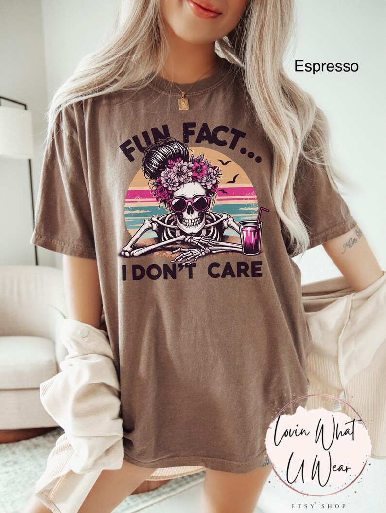 May include: Brown t-shirt with a graphic of a skeleton with a flower crown and sunglasses holding a glass of pink liquid. The text "Fun Fact... I don't care" is printed on the shirt.