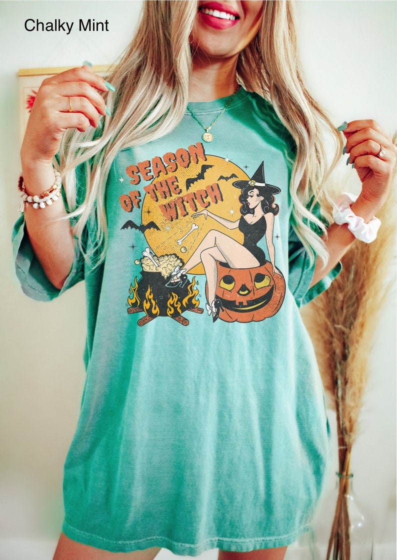 Puede incluir: Una camiseta azul turquesa con un gr&aacute;fico de estilo vintage de una bruja sentada en una calabaza de Halloween. El gr&aacute;fico dice "Season of the Witch".