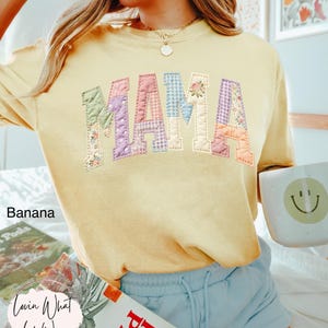 Puede incluir: Una sudadera amarilla con la palabra "MAMA" en letras de patchwork. Las letras est&aacute;n hechas de varias telas florales y estampadas. La palabra "Banana" est&aacute; impresa en el lado izquierdo. Una taza con una carita sonriente se sostiene en la mano derecha.