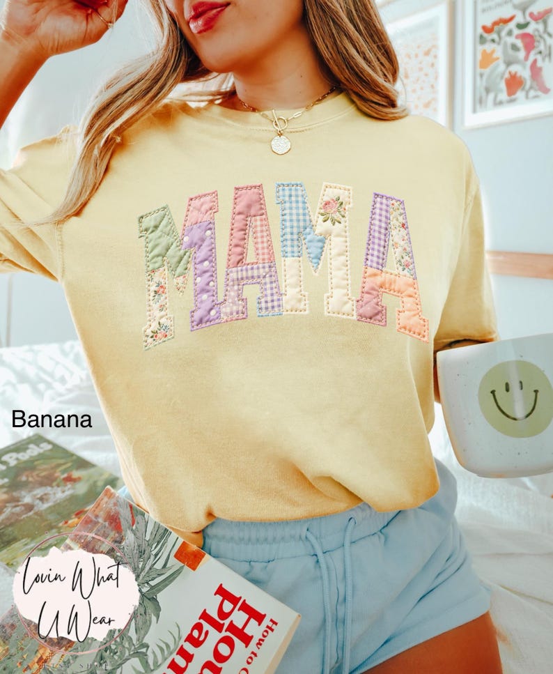 Puede incluir: Una sudadera amarilla con la palabra "MAMA" en letras de patchwork. Las letras est&aacute;n hechas de varias telas florales y estampadas. La palabra "Banana" est&aacute; impresa en el lado izquierdo. Una taza con una carita sonriente se sostiene en la mano derecha.